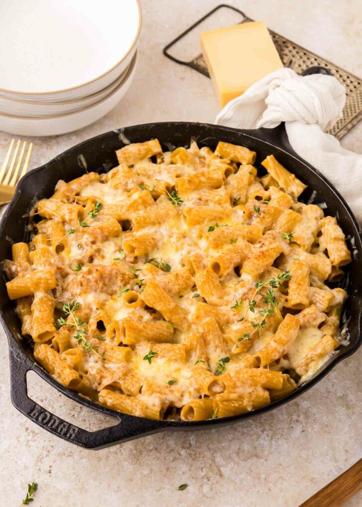 A cast iron skillet filled with rigatoni pasta with caramelized onions topped with melted cheese and fresh herbs, sitting on a light countertop next to plates, a block of cheese, and a white napkin.