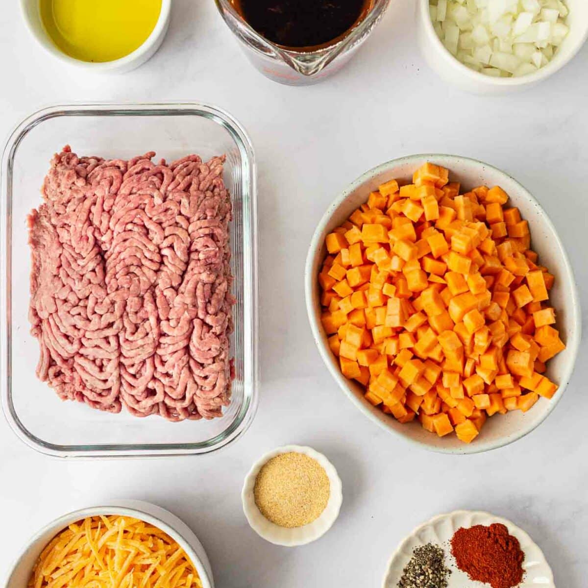 Ingredients on a white surface, including ground beef, cubed sweet potatoes, shredded cheese, chopped onions, olive oil, spices, garlic powder, and a cup of beef broth