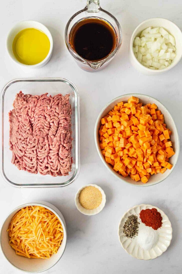 Ingredients on a white surface, including ground beef, cubed sweet potatoes, shredded cheese, chopped onions, olive oil, spices, garlic powder, and a cup of beef broth