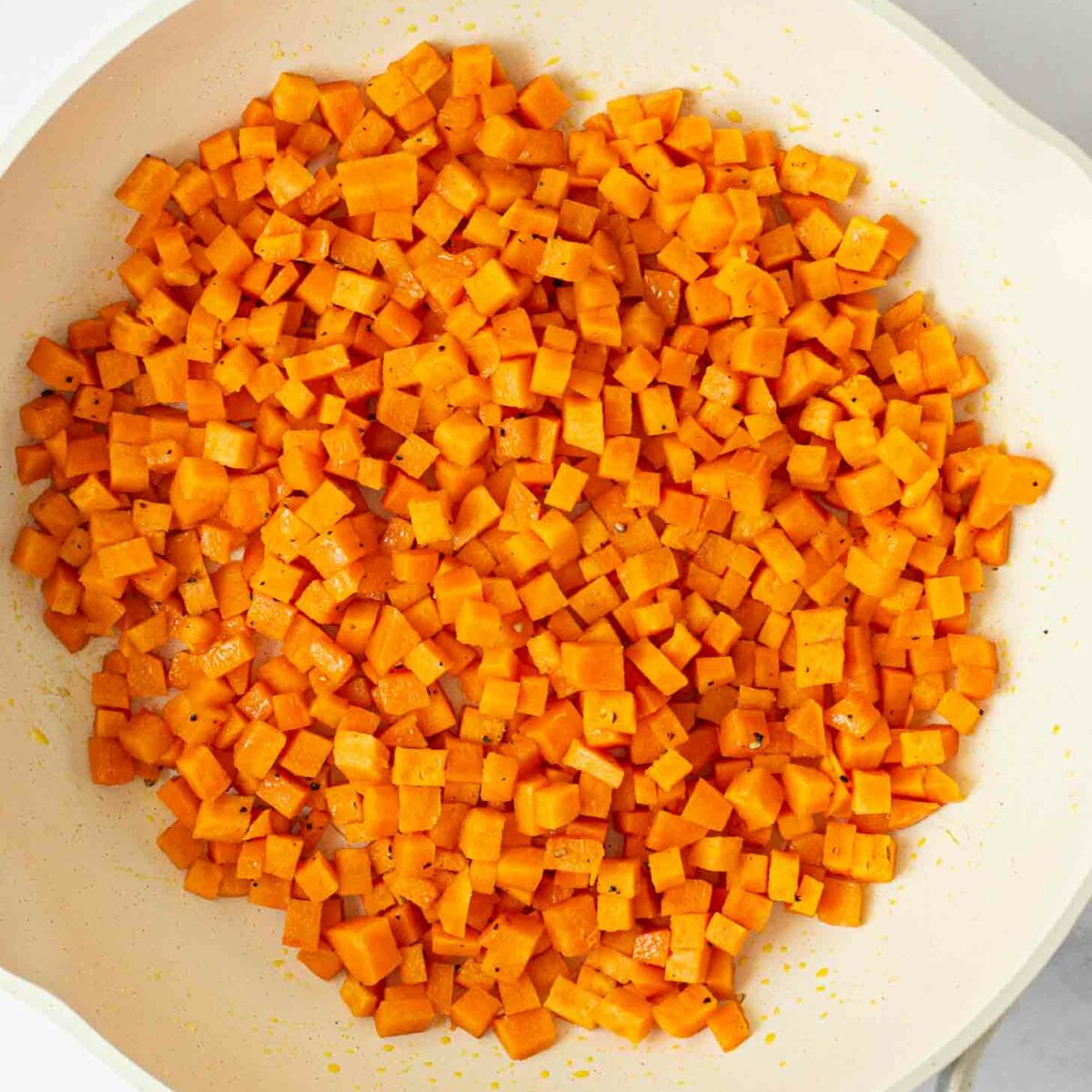 Diced sweet potatoes sautéing in a white frying pan, lightly seasoned and cooked on a white surface.