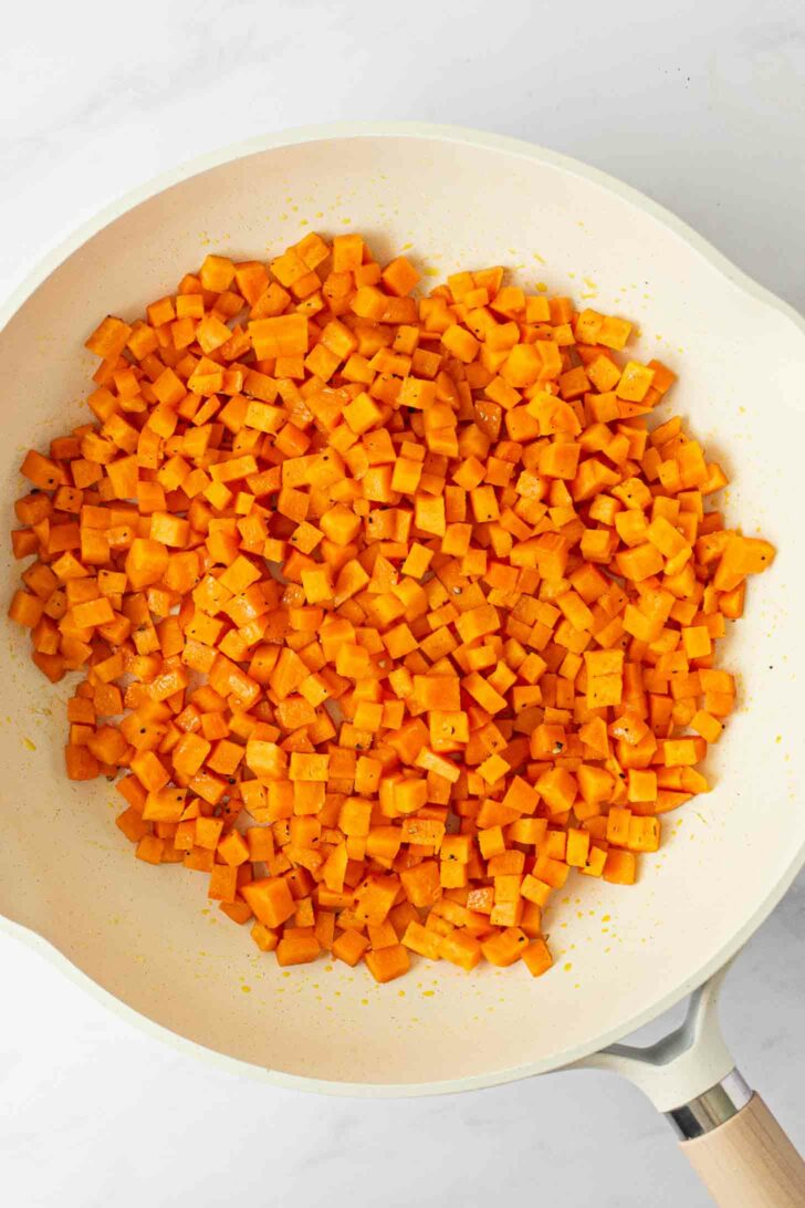Diced sweet potatoes sautéing in a white frying pan, lightly seasoned and cooked on a white surface.