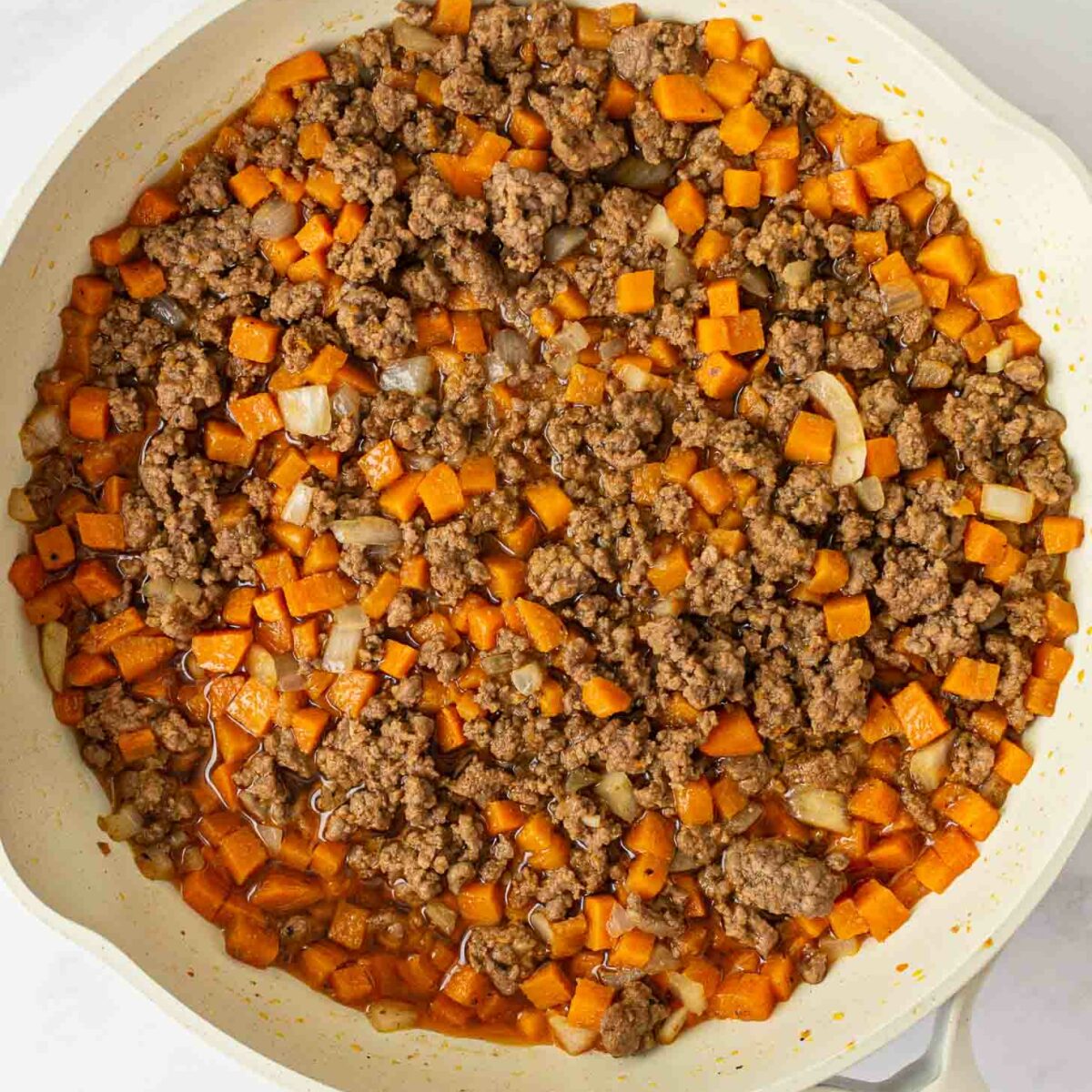 A white skillet filled with cooked ground beef, diced orange sweet potatoes, and onions, all mixed together on a white surface.