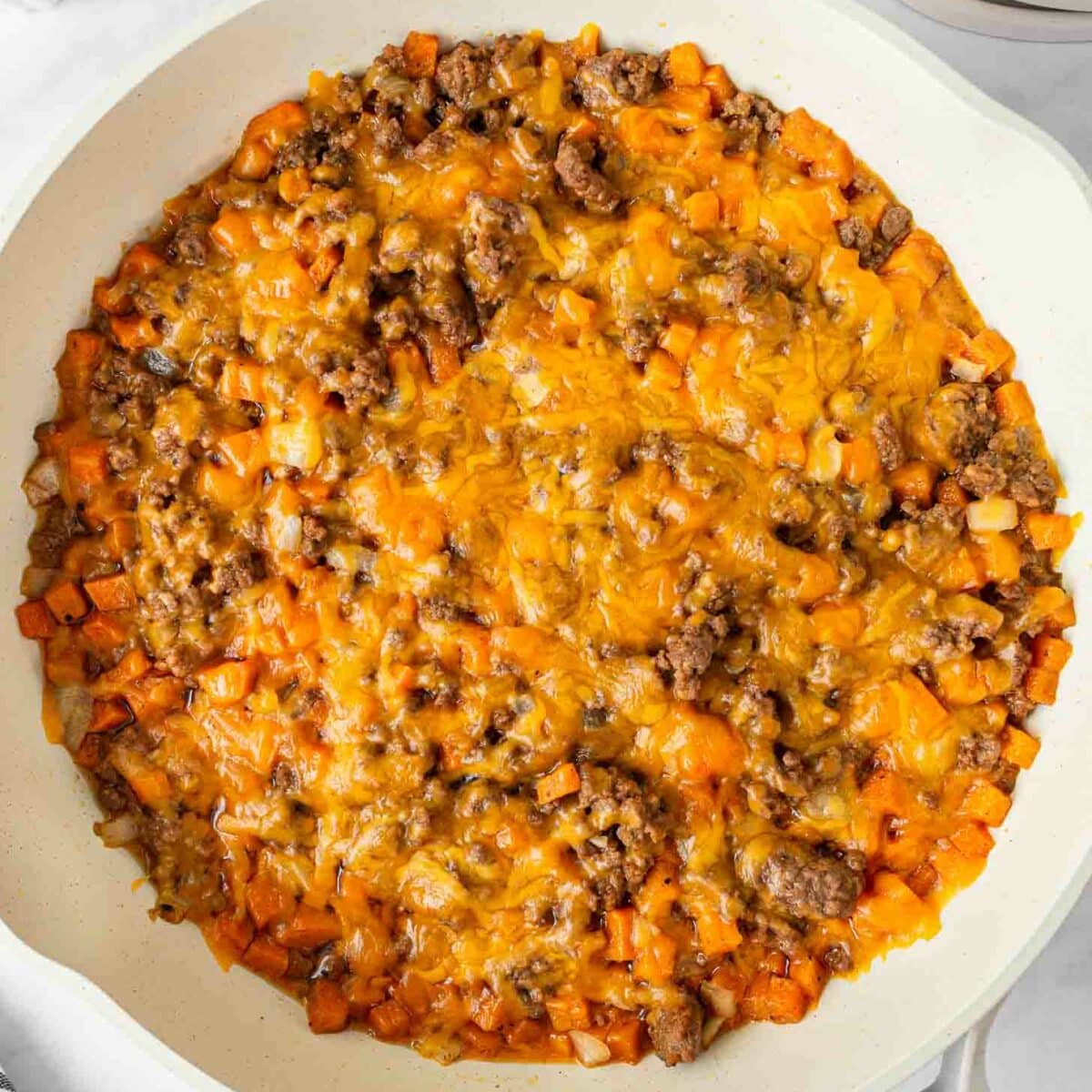 A skillet filled with a cooked mixture of ground beef, diced sweet potatoes, onions, and melted cheddar cheese on top, with a striped napkin, seasonings, and utensils nearby.