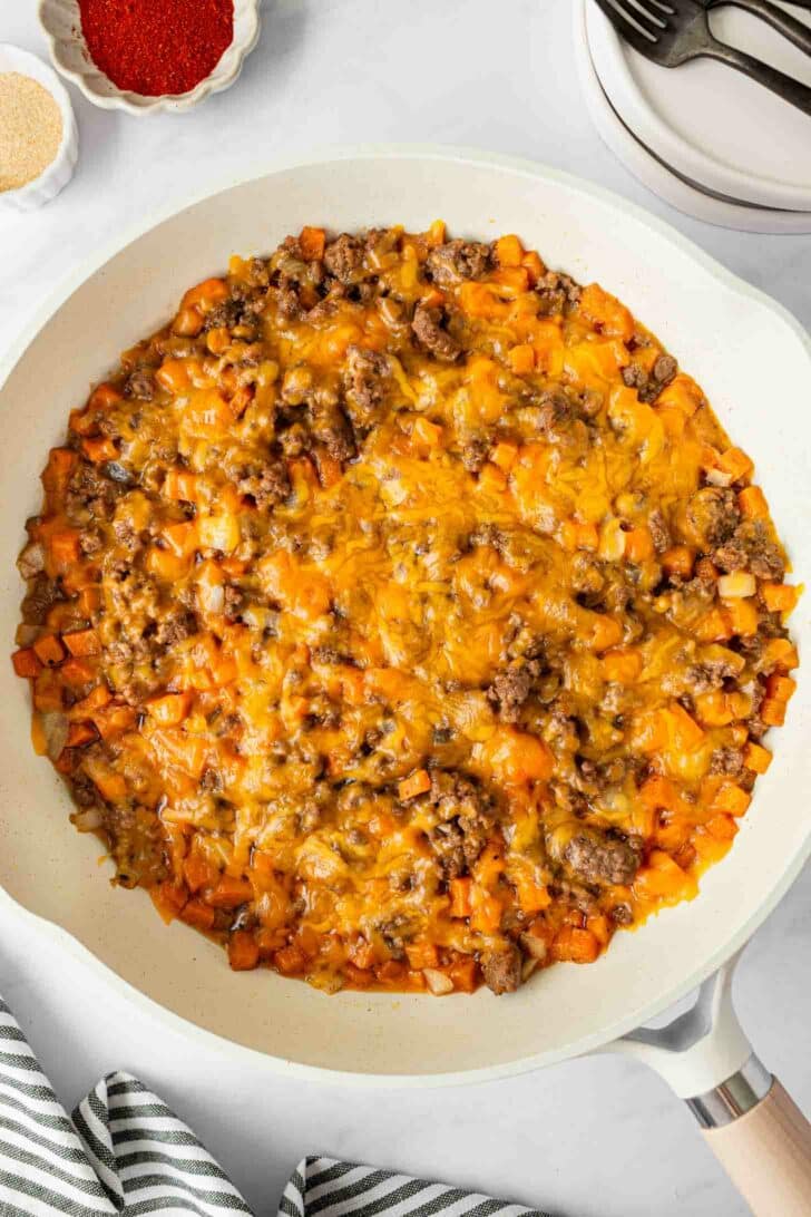 A skillet filled with a cooked mixture of ground beef, diced sweet potatoes, onions, and melted cheddar cheese on top, with a striped napkin, seasonings, and utensils nearby.