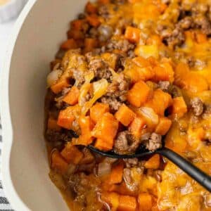 A close-up of a casserole dish filled with a cheesy mixture of ground beef, sweet potatoes, and onions. A serving spoon is scooping out the hearty, melted cheese-topped casserole.
