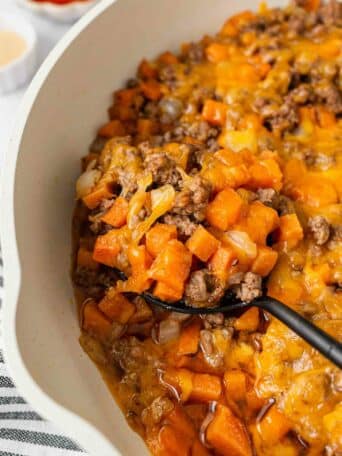 A close-up of a casserole dish filled with a cheesy mixture of ground beef, sweet potatoes, and onions. A serving spoon is scooping out the hearty, melted cheese-topped casserole.