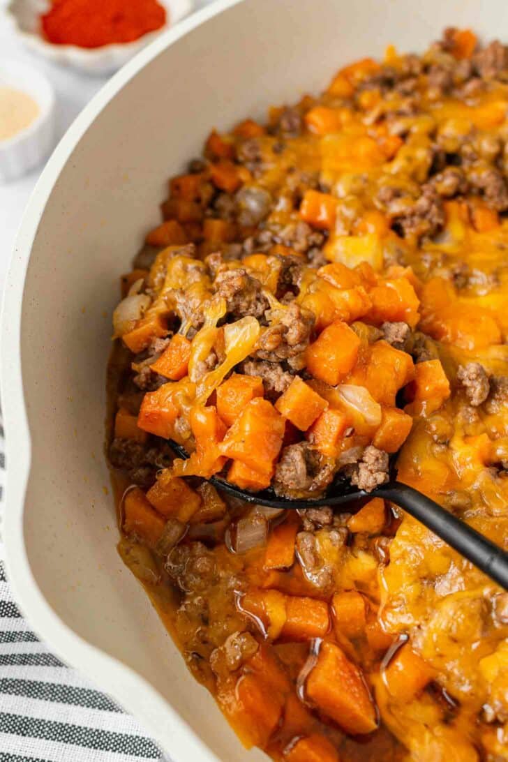A close-up of a casserole dish filled with a cheesy mixture of ground beef, sweet potatoes, and onions. A serving spoon is scooping out the hearty, melted cheese-topped casserole.