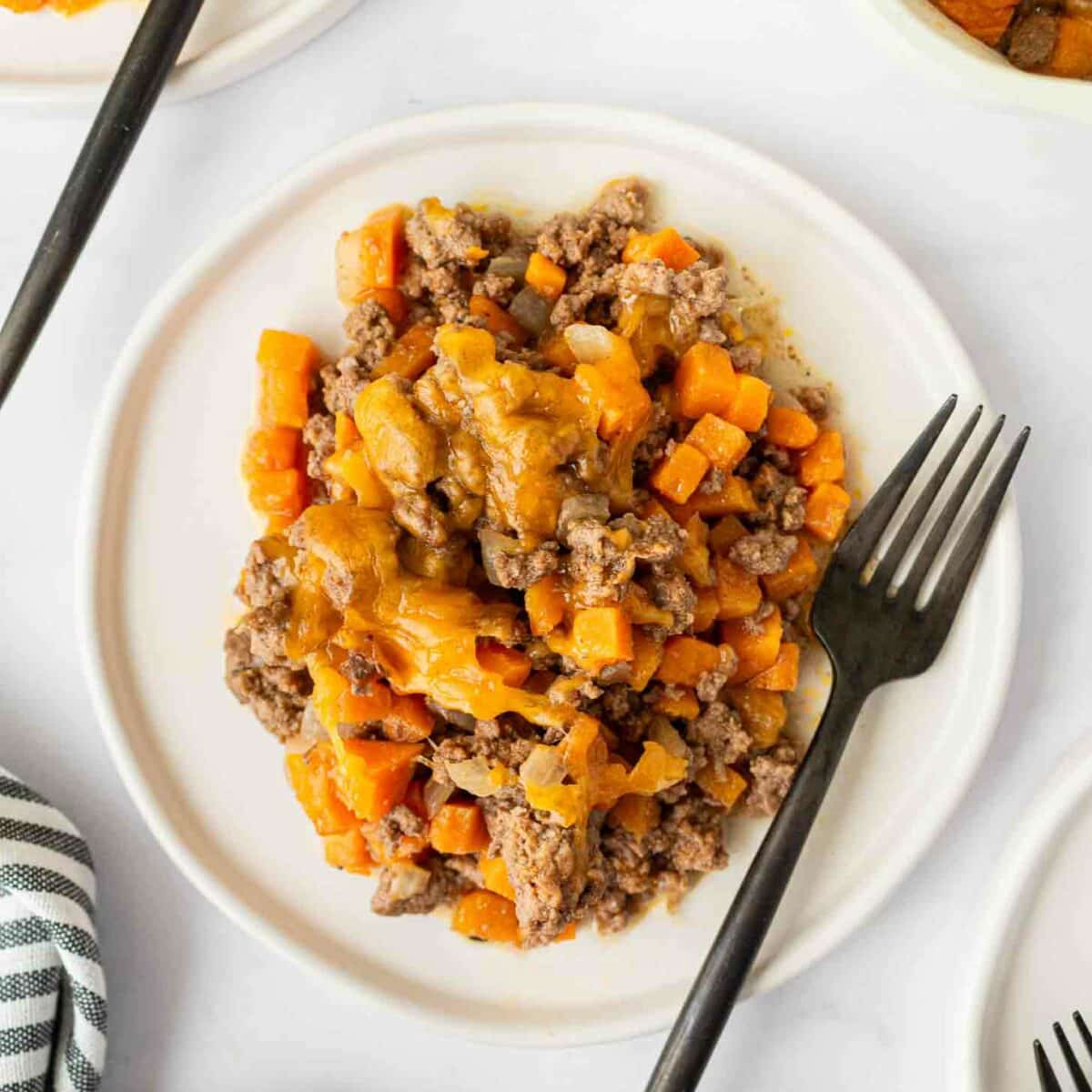 A white plate with a serving of ground beef, diced sweet potatoes, and melted cheese, with a black fork on the side; a striped cloth is partially visible in the corner.