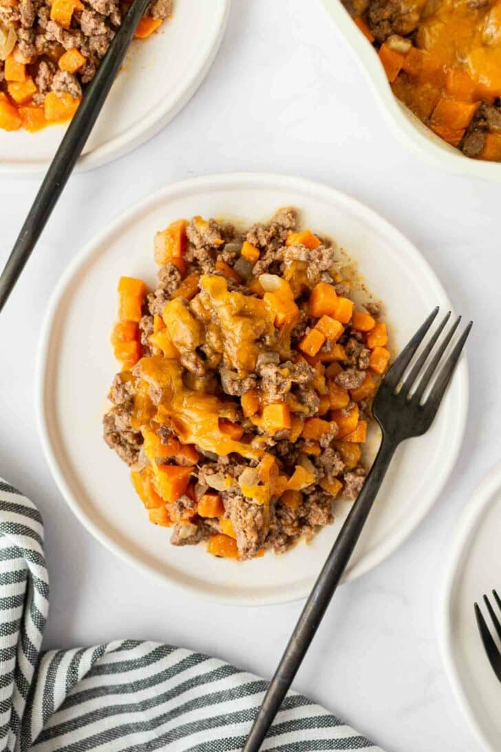A white plate with a serving of ground beef, diced sweet potatoes, and melted cheese, with a black fork on the side; a striped cloth is partially visible in the corner.