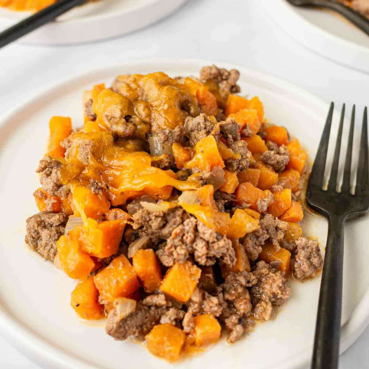 A serving of ground beef and diced sweet potatoes topped with melted cheese on a white plate, with a black fork beside it. Two similar plates are visible in the background.