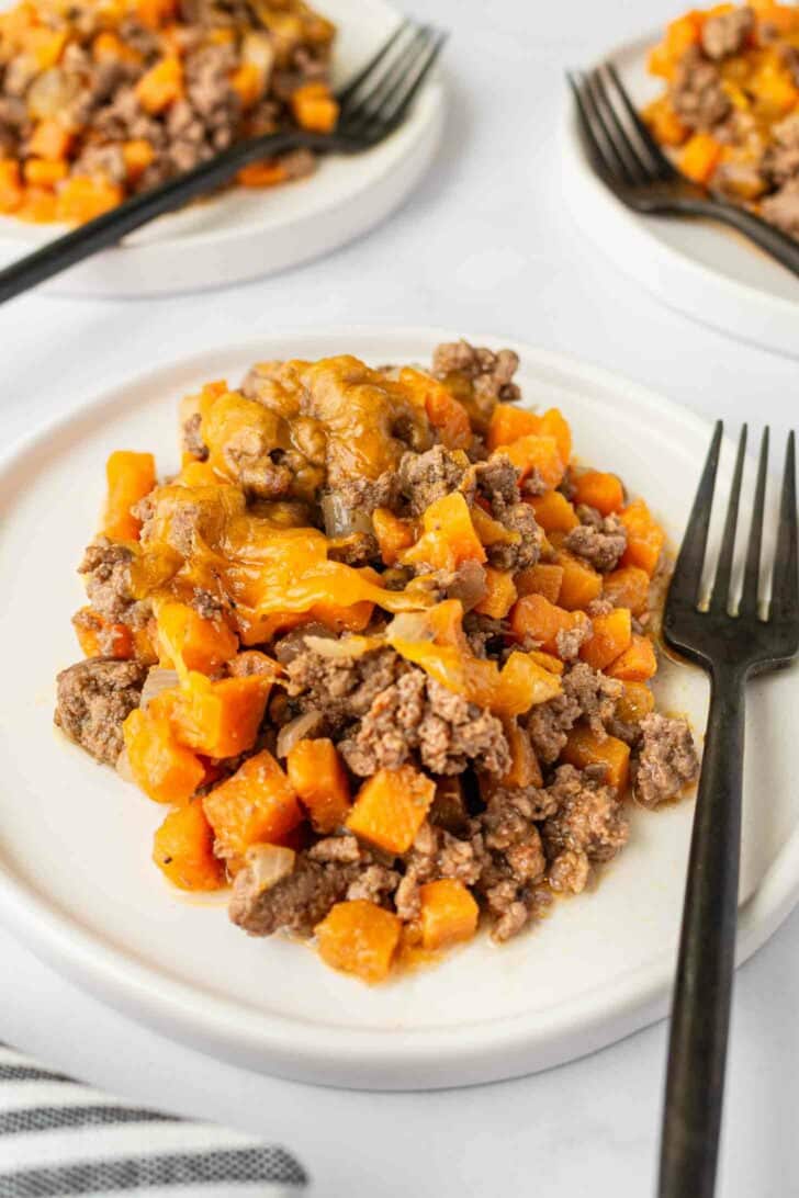 A serving of ground beef and diced sweet potatoes topped with melted cheese on a white plate, with a black fork beside it. Two similar plates are visible in the background.