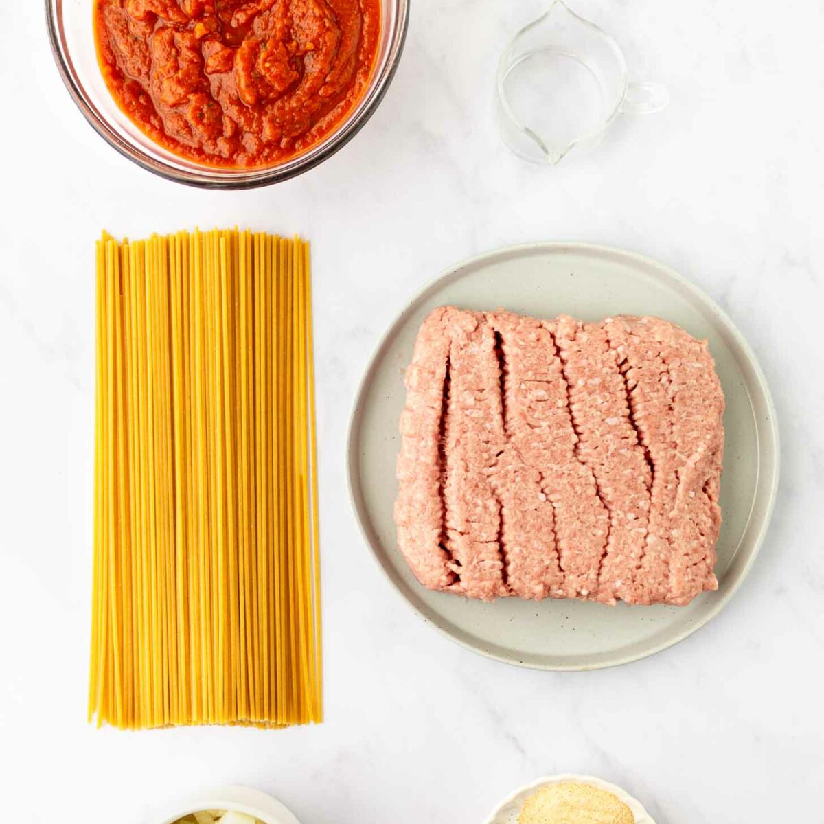 Ingredients for a pasta dish arranged on a white surface: uncooked spaghetti, ground meat on a plate, a bowl of tomato sauce, diced onions, mixed dried spices, and a small bowl of water.