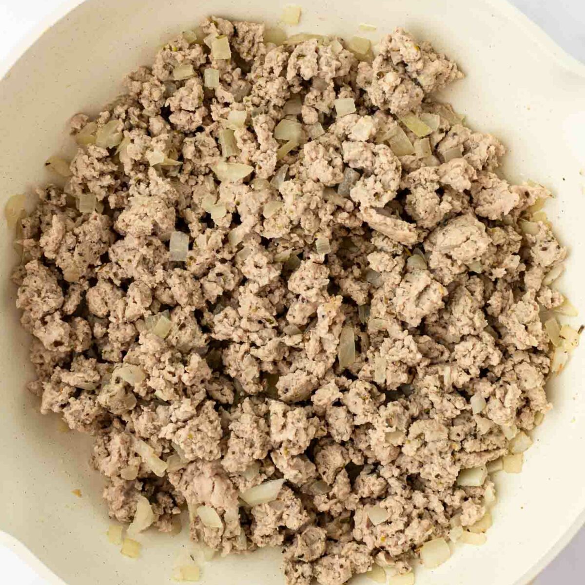 Cooked ground turkey with small pieces of sautéed onion in a white frying pan, viewed from above on a light background.