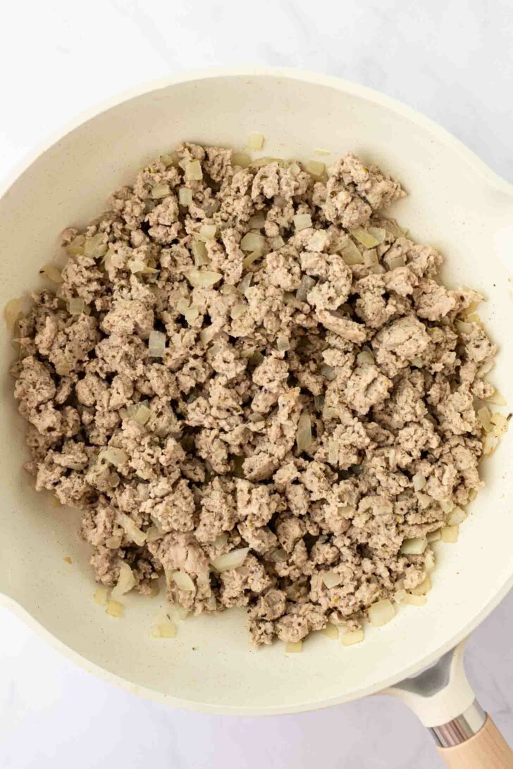 Cooked ground turkey with small pieces of sautéed onion in a white frying pan, viewed from above on a light background.