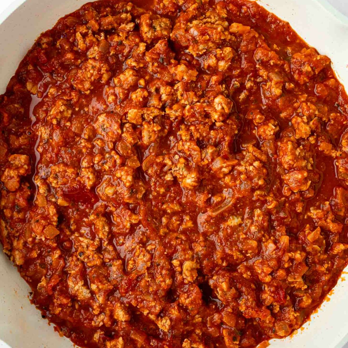 A white skillet filled with cooked ground turkey in a rich, red tomato-based sauce, resembling a homemade meat sauce, set against a white background.