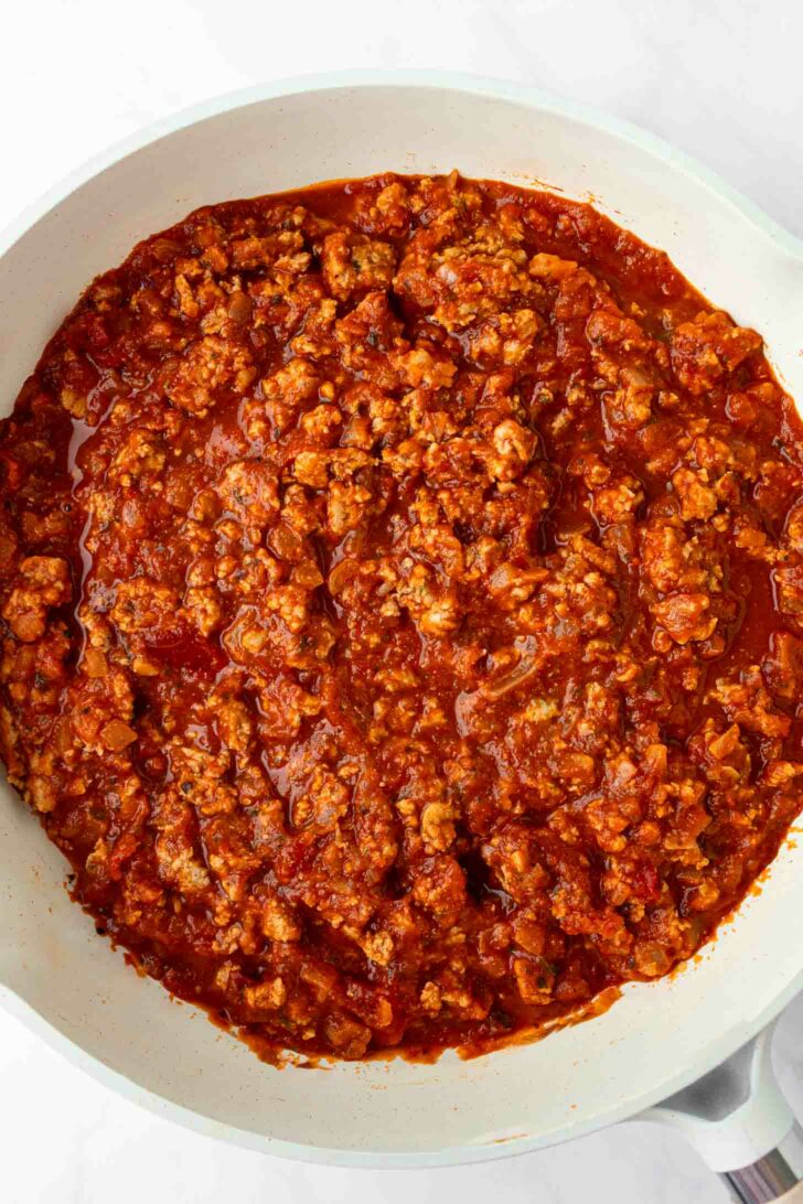 A white skillet filled with cooked ground turkey in a rich, red tomato-based sauce, resembling a homemade meat sauce, set against a white background.