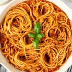 A skillet filled with spaghetti pasta mixed with meat sauce, garnished with a small cluster of fresh basil leaves in the center. Plates, utensils, and a bowl of chopped herbs are nearby on a white surface.