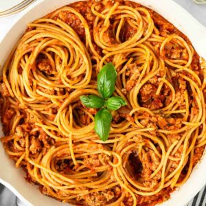 A skillet filled with spaghetti pasta mixed with meat sauce, garnished with a small cluster of fresh basil leaves in the center. Plates, utensils, and a bowl of chopped herbs are nearby on a white surface.