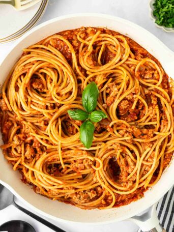 A skillet filled with spaghetti pasta mixed with meat sauce, garnished with a small cluster of fresh basil leaves in the center. Plates, utensils, and a bowl of chopped herbs are nearby on a white surface.