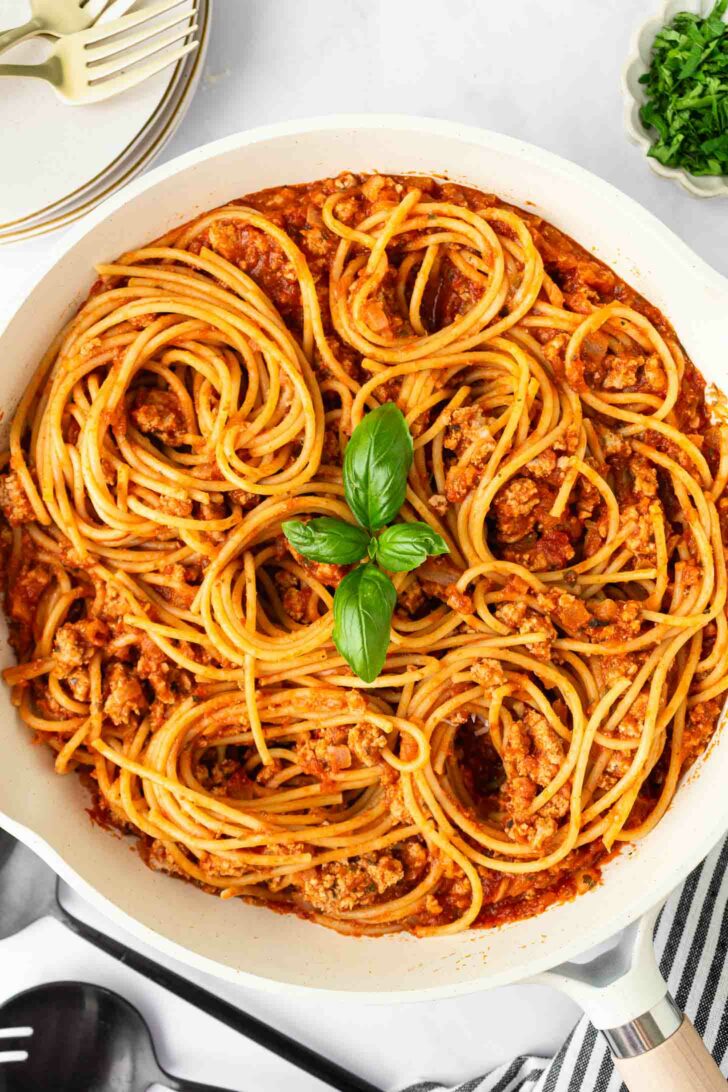 A skillet filled with spaghetti pasta mixed with meat sauce, garnished with a small cluster of fresh basil leaves in the center. Plates, utensils, and a bowl of chopped herbs are nearby on a white surface.