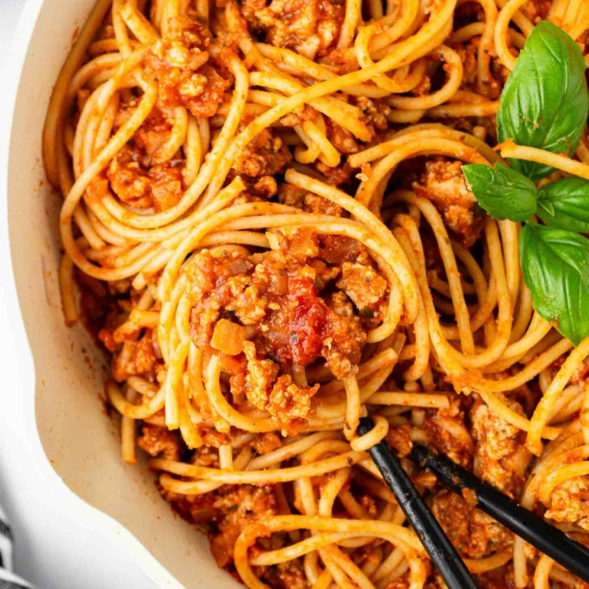 A close-up of spaghetti with meat sauce in a white pan, garnished with fresh basil. Black spoons are holding up a twirl of pasta. A striped cloth is partially visible in the corner.