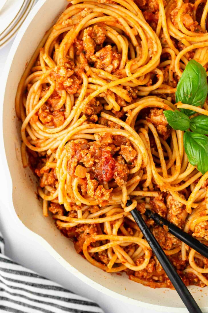 A close-up of spaghetti with meat sauce in a white pan, garnished with fresh basil. Black spoons are holding up a twirl of pasta. A striped cloth is partially visible in the corner.