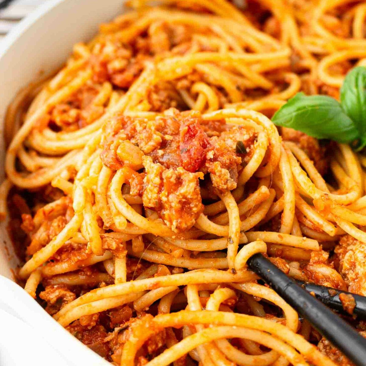 Close-up of spaghetti coated in a chunky tomato and meat sauce, served in a white bowl, with a black serving spoon and a sprig of fresh basil for garnish.