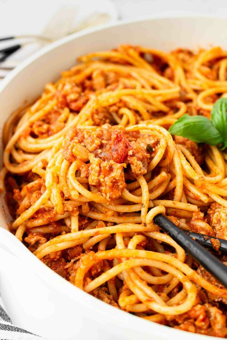 Close-up of spaghetti coated in a chunky tomato and meat sauce, served in a white bowl, with a black serving spoon and a sprig of fresh basil for garnish.