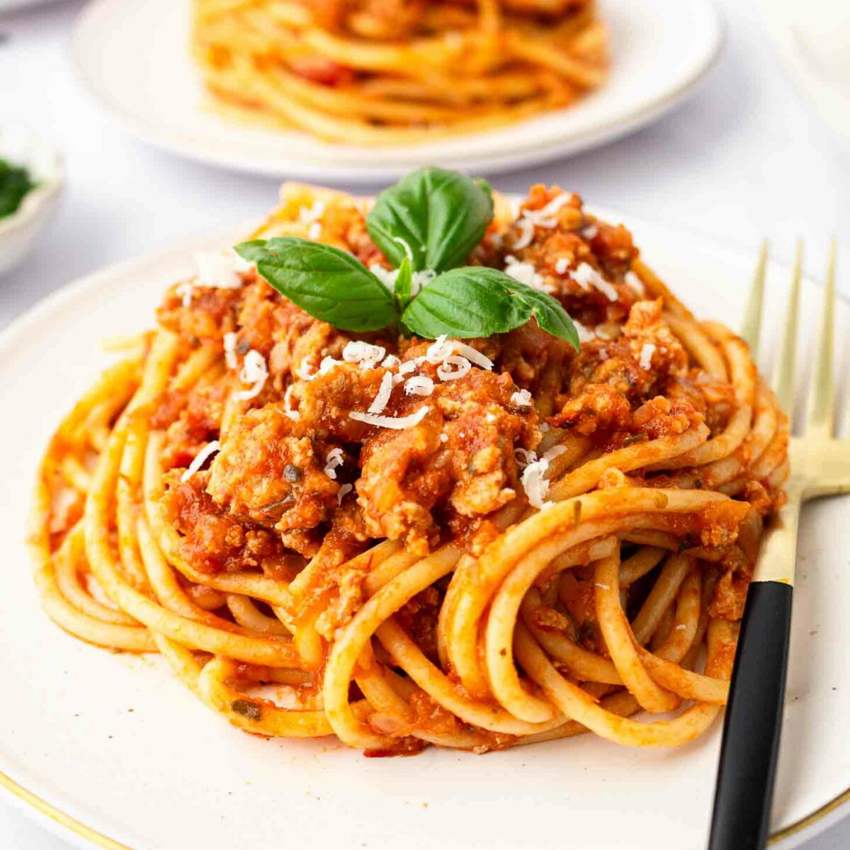 A plate of spaghetti topped with ground turkey, grated cheese, and fresh basil, with a black and gold fork on the side. Another plate of pasta is visible in the background.