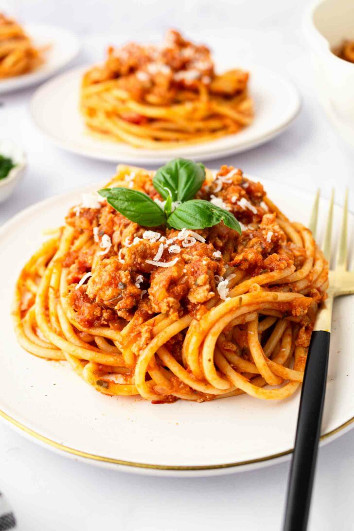 A plate of spaghetti topped with ground turkey, grated cheese, and fresh basil, with a black and gold fork on the side. Another plate of pasta is visible in the background.