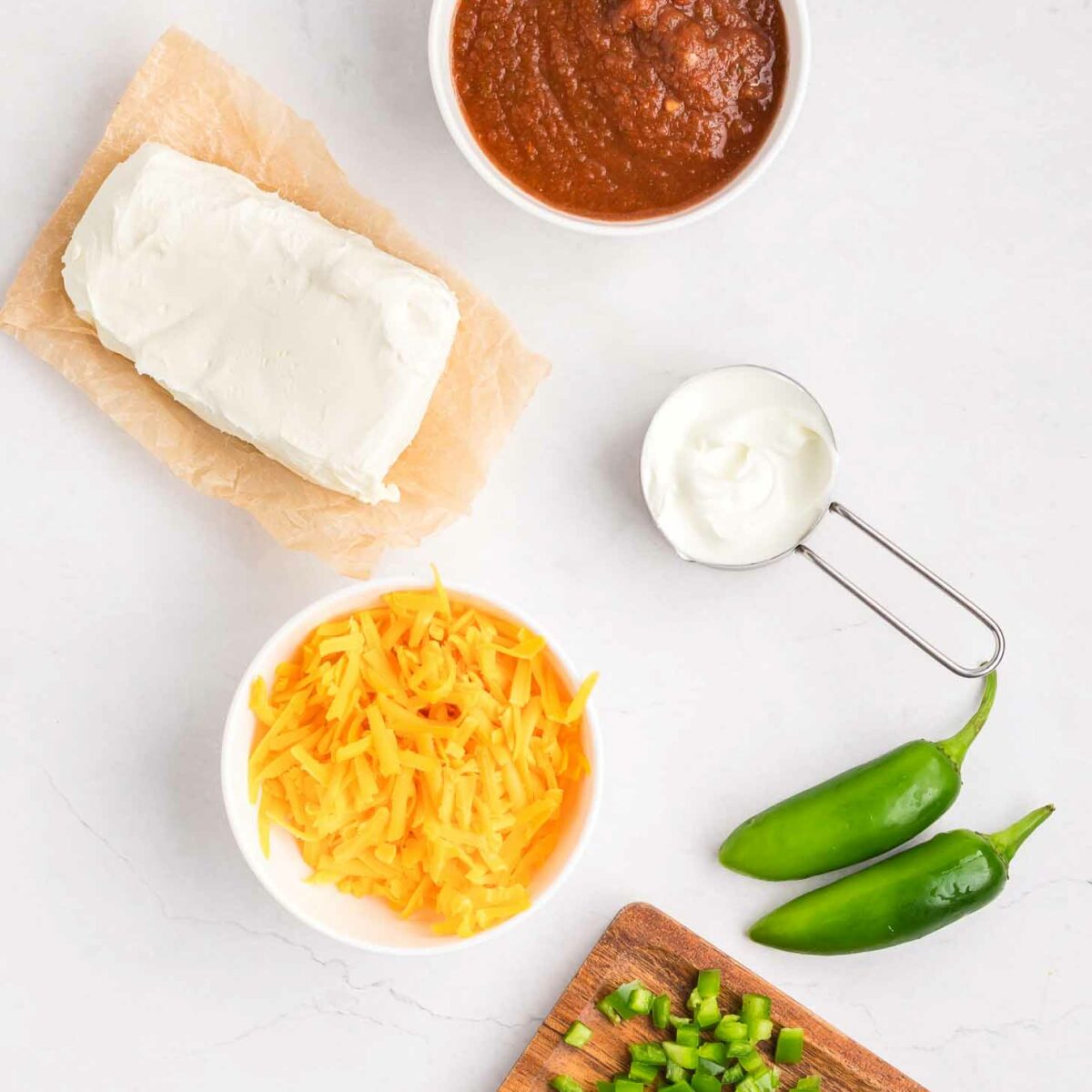 Ingredients on a white surface: a block of cream cheese on parchment, a bowl of salsa, a small cup of sour cream, shredded cheddar cheese in a bowl, two whole jalapeños, and chopped jalapeños on a cutting board with a knife.