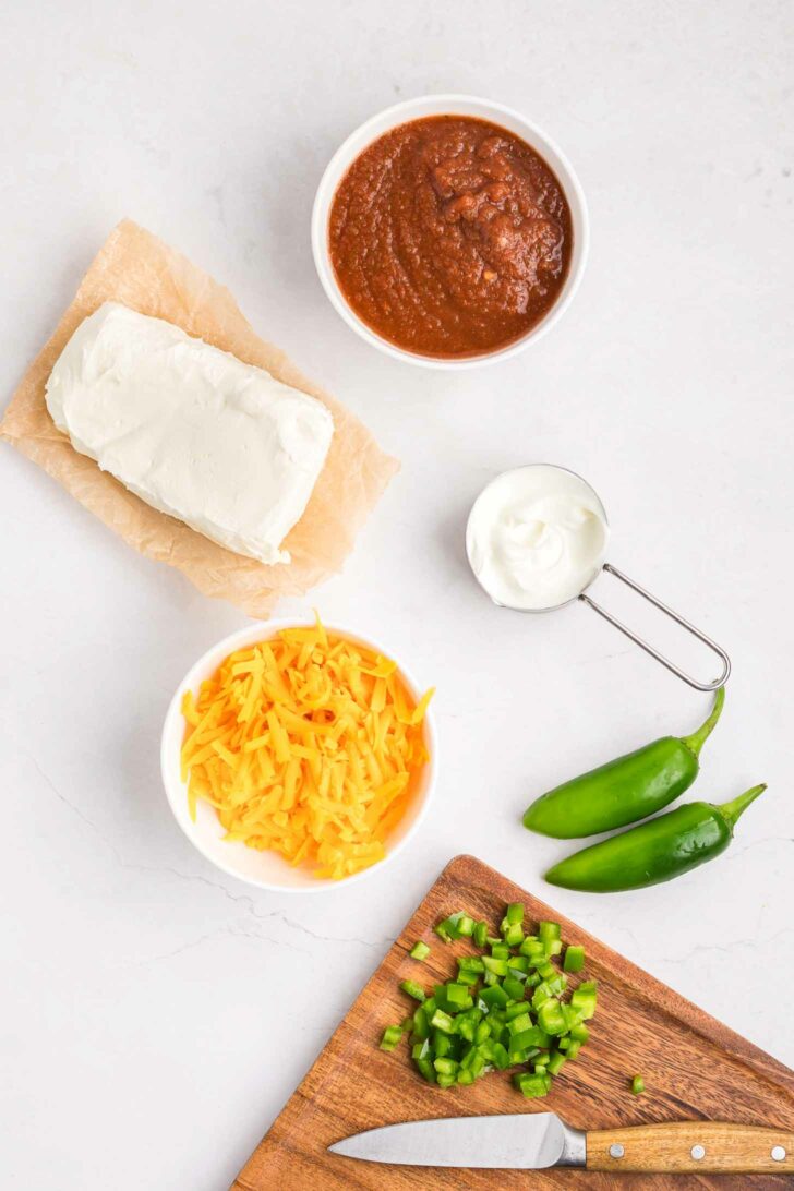 Ingredients on a white surface: a block of cream cheese on parchment, a bowl of salsa, a small cup of sour cream, shredded cheddar cheese in a bowl, two whole jalapeños, and chopped jalapeños on a cutting board with a knife.