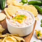 A white bowl of creamy dip topped with shredded cheese and chopped jalapeños is surrounded by ridged potato chips on a wooden board, with fresh jalapeños scattered nearby.