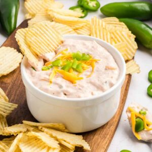 A white bowl of creamy dip topped with shredded cheese and chopped jalapeños is surrounded by ridged potato chips on a wooden board, with fresh jalapeños scattered nearby.