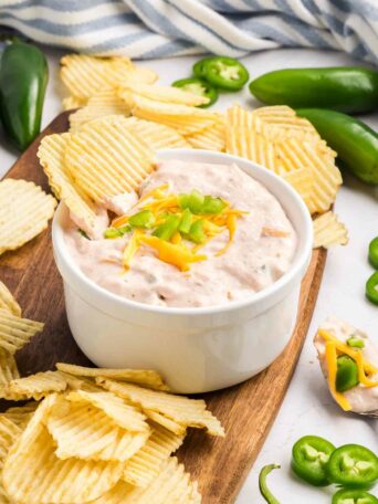 A white bowl of creamy dip topped with shredded cheese and chopped jalapeños is surrounded by ridged potato chips on a wooden board, with fresh jalapeños scattered nearby.