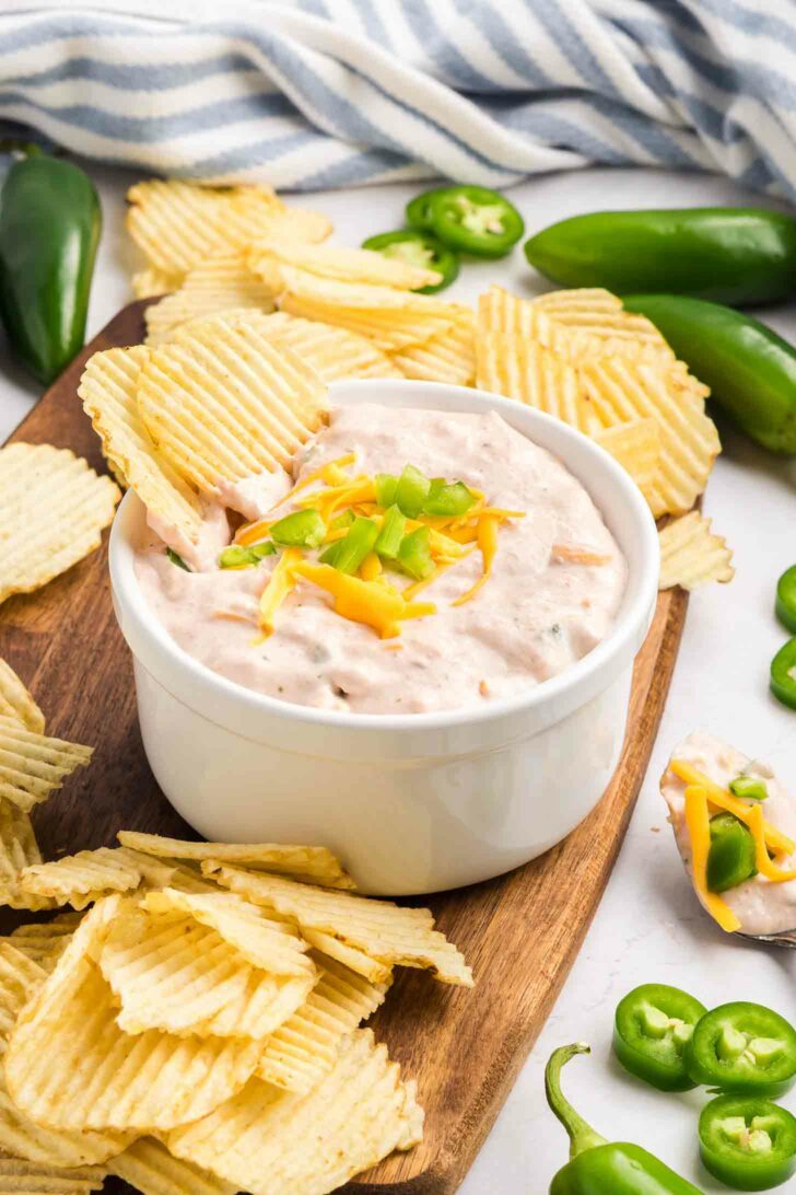 A white bowl of creamy dip topped with shredded cheese and chopped jalapeños is surrounded by ridged potato chips on a wooden board, with fresh jalapeños scattered nearby.