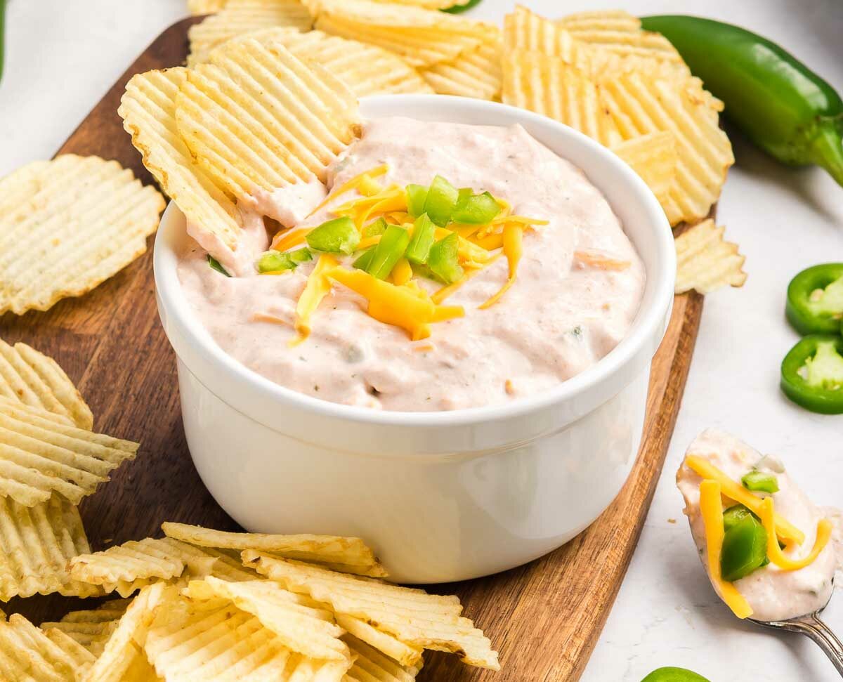A white bowl of creamy dip topped with shredded cheddar cheese and chopped jalapeños, surrounded by ridged potato chips on a wooden board, with jalapeño slices scattered nearby.