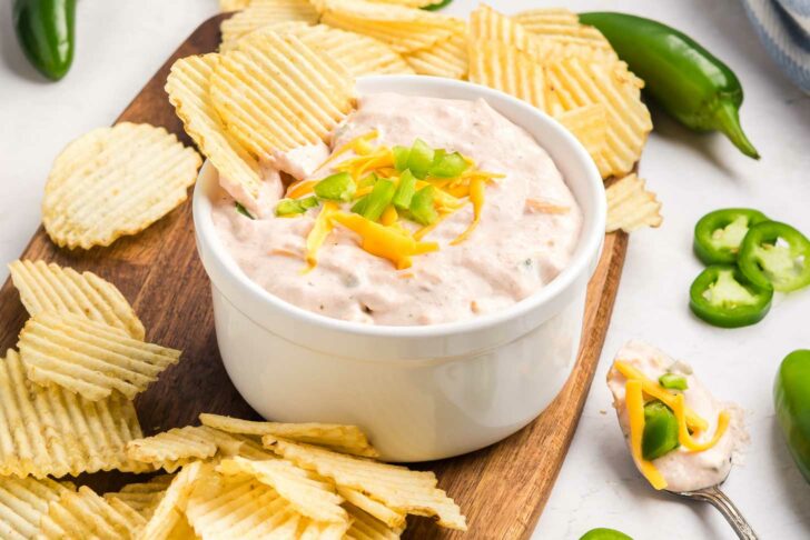 A white bowl of creamy dip topped with shredded cheddar cheese and chopped jalapeños, surrounded by ridged potato chips on a wooden board, with jalapeño slices scattered nearby.