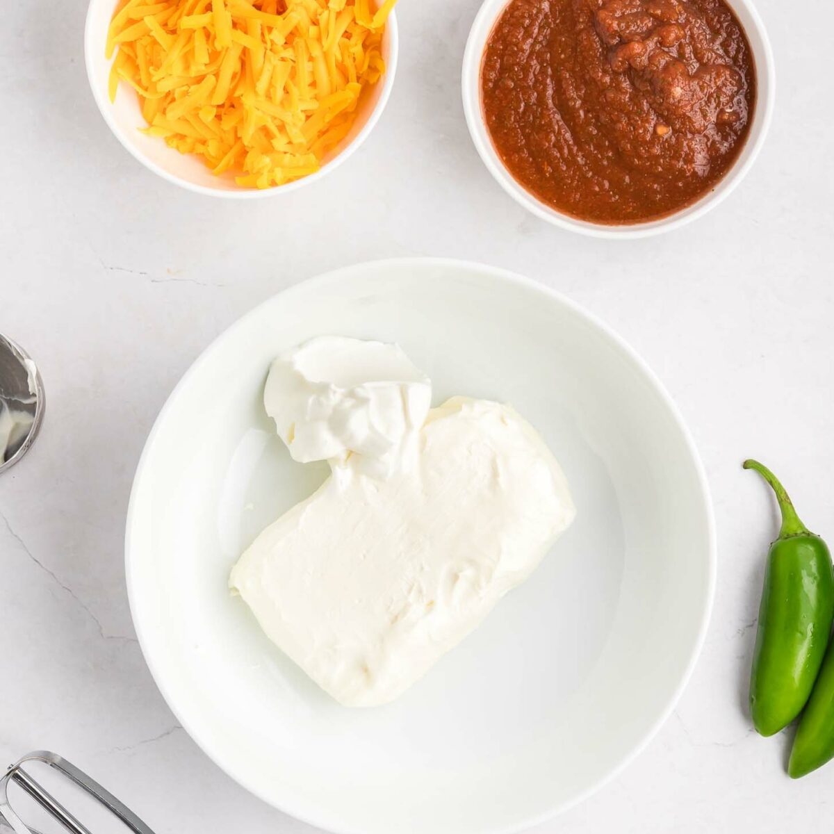A bowl with cream cheese and sour cream, a bowl of shredded cheddar cheese, a bowl of salsa, a hand mixer beater, and two jalapeños on a white countertop.