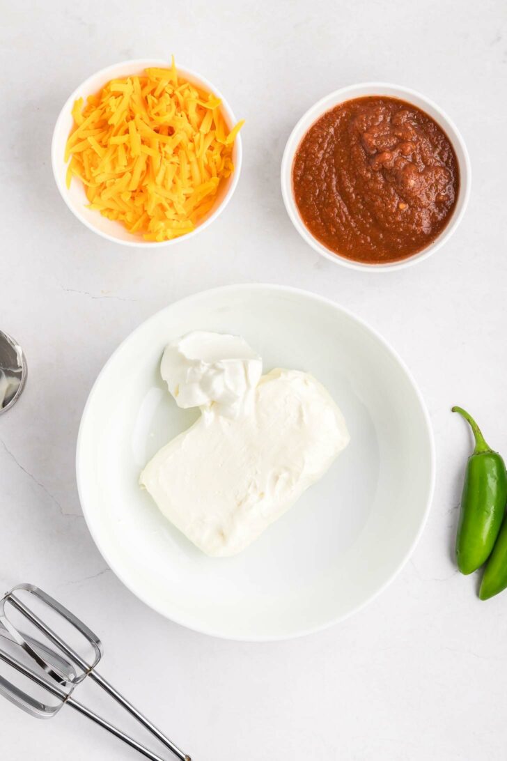 A bowl with cream cheese and sour cream, a bowl of shredded cheddar cheese, a bowl of salsa, a hand mixer beater, and two jalapeños on a white countertop.