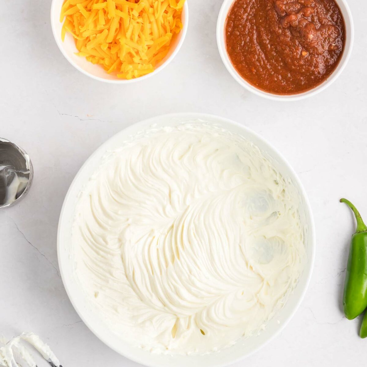 A bowl of whipped cream cheese and sour cream sits beside a small bowl of shredded cheddar cheese and a bowl of salsa. Part of a hand mixer and beaters are visible, along with a jalapeño on a white surface.