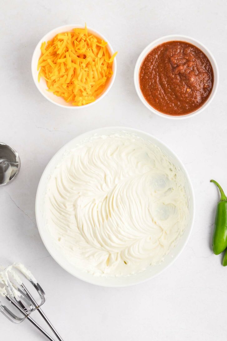 A bowl of whipped cream cheese and sour cream sits beside a small bowl of shredded cheddar cheese and a bowl of salsa. Part of a hand mixer and beaters are visible, along with a jalapeño on a white surface.