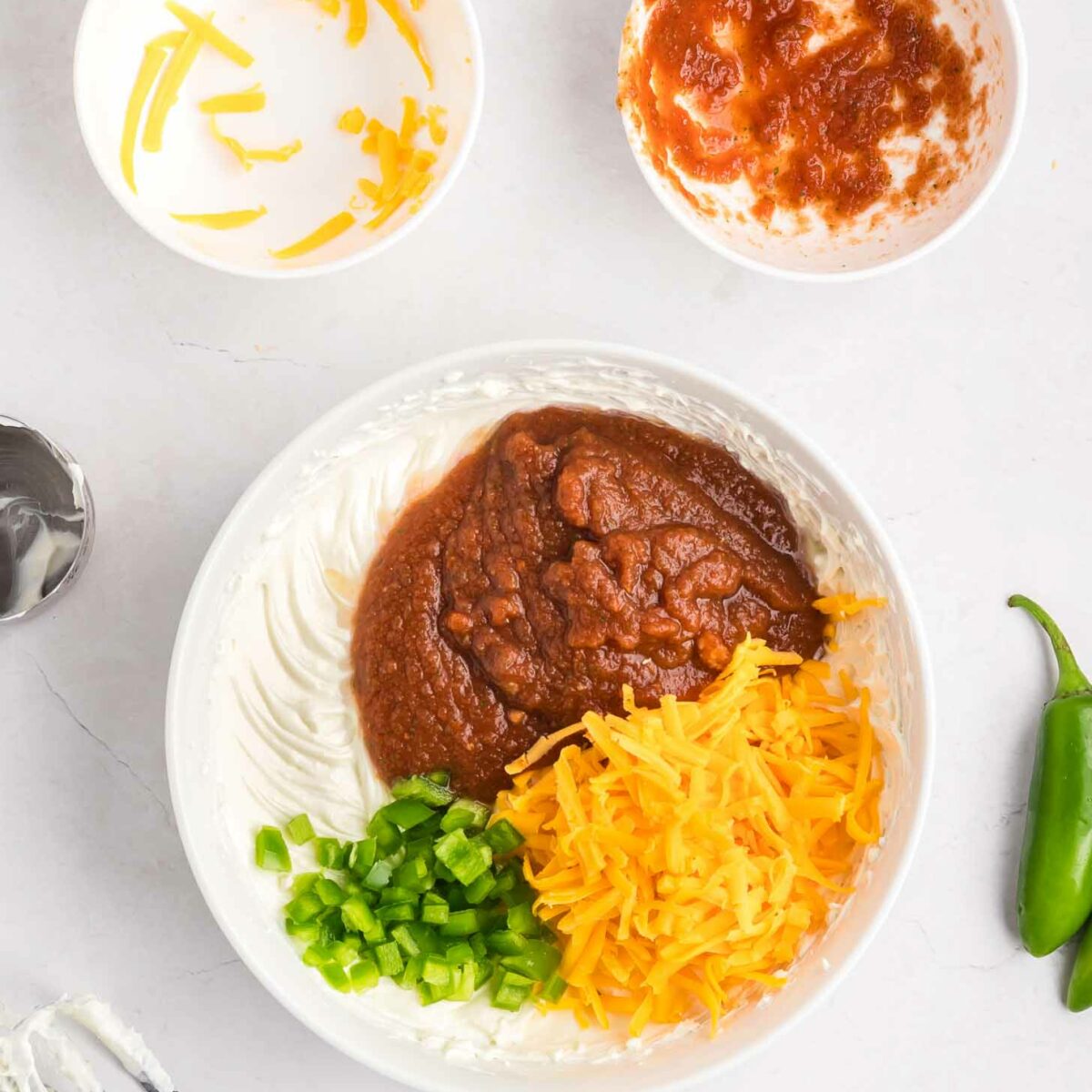 A bowl with cream cheese, sour cream, salsa, shredded cheddar cheese, and chopped jalapeños, ready to be mixed. Nearby are bowls with leftover cheese and salsa, a mixer with beaters, and a fresh jalapeño pepper.