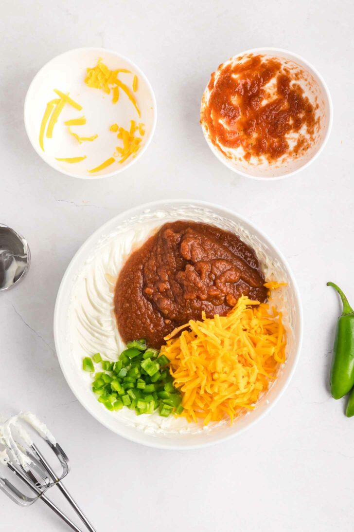 A bowl with cream cheese, sour cream, salsa, shredded cheddar cheese, and chopped jalapeños, ready to be mixed. Nearby are bowls with leftover cheese and salsa, a mixer with beaters, and a fresh jalapeño pepper.