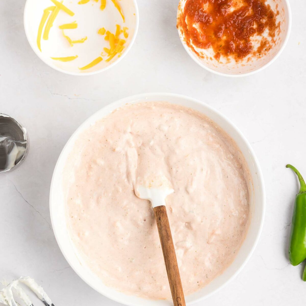 A mixing bowl with a creamy pale pink dip, a spatula, a bowl with shredded cheese, a bowl with salsa, a metal mixer, and a jalapeño on a white surface.