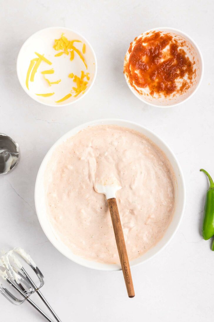 A mixing bowl with a creamy pale pink dip, a spatula, a bowl with shredded cheese, a bowl with salsa, a metal mixer, and a jalapeño on a white surface.
