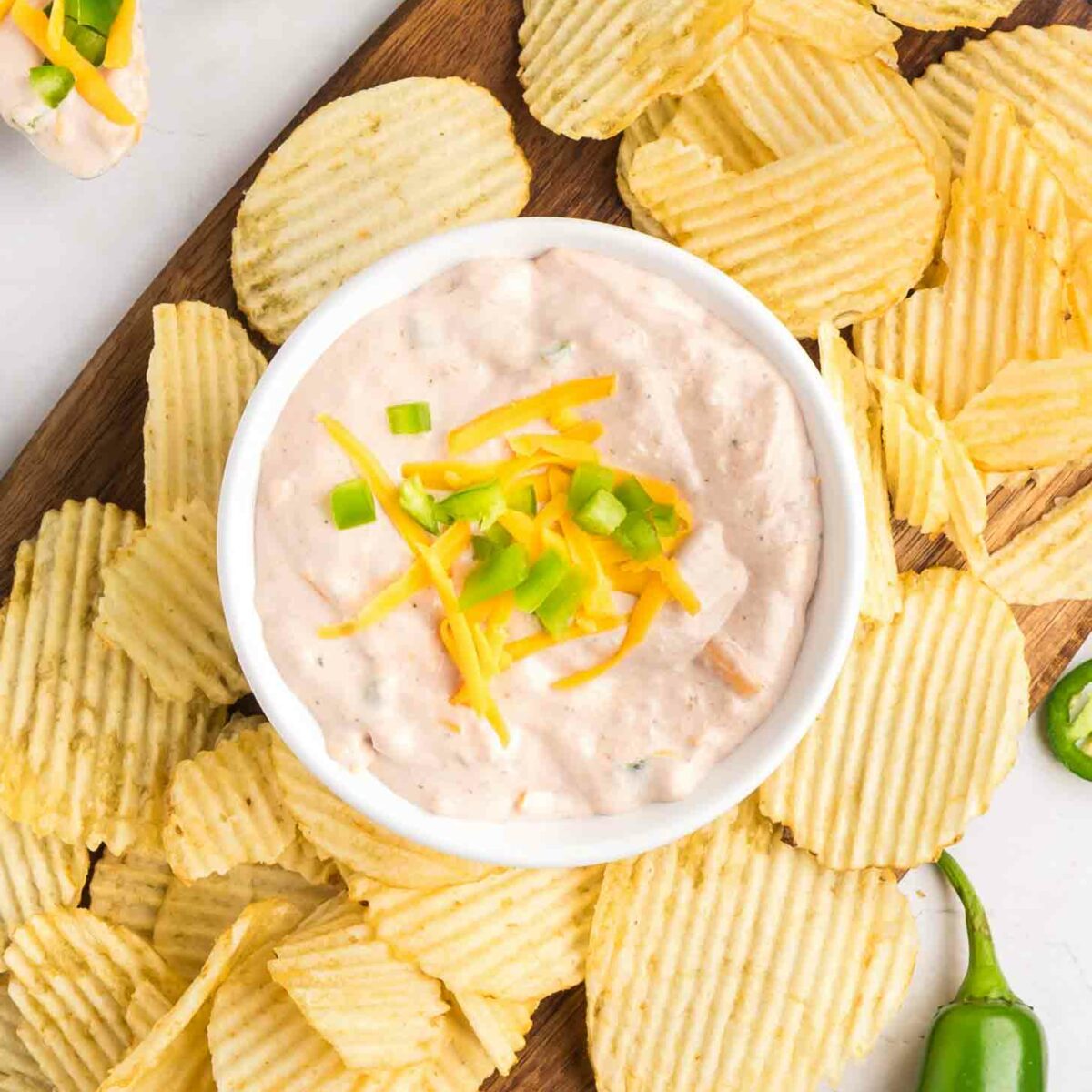 A bowl of creamy dip topped with shredded cheese and diced green onions is surrounded by ridged potato chips on a wooden board, with whole and sliced jalapeños nearby.