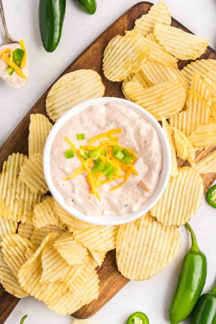 A bowl of creamy dip topped with shredded cheese and diced green onions is surrounded by ridged potato chips on a wooden board, with whole and sliced jalapeños nearby.