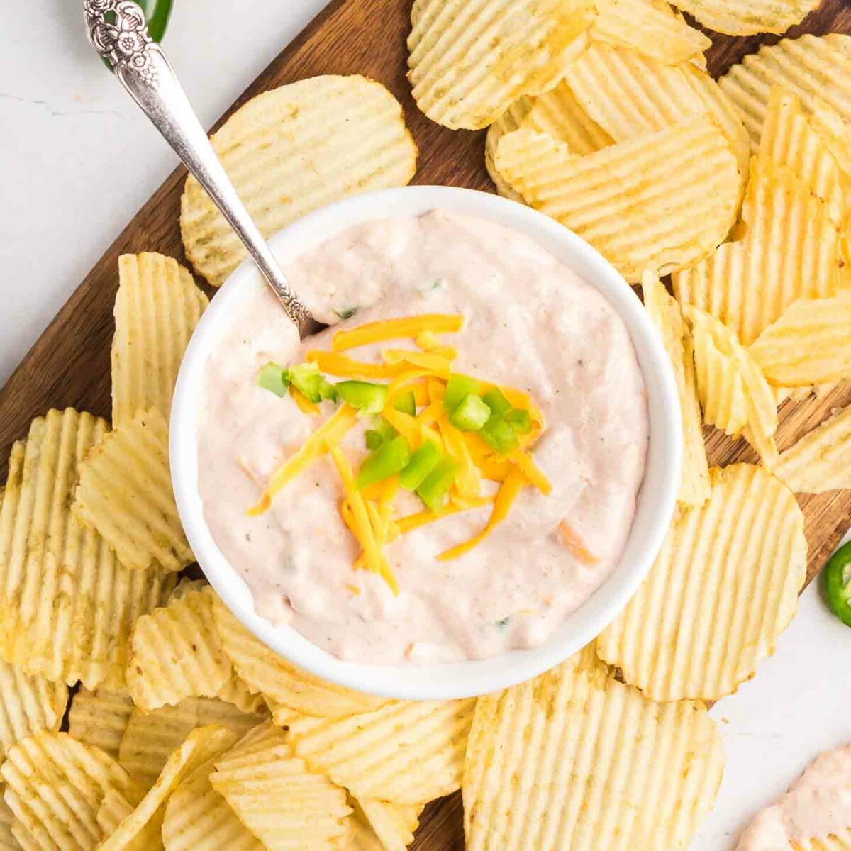 A bowl of creamy dip topped with shredded cheddar cheese and chopped green onion is surrounded by crinkle-cut potato chips on a wooden board, with jalapeño peppers nearby.