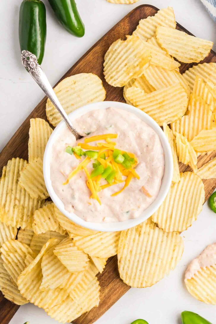 A bowl of creamy dip topped with shredded cheddar cheese and chopped green onion is surrounded by crinkle-cut potato chips on a wooden board, with jalapeño peppers nearby.